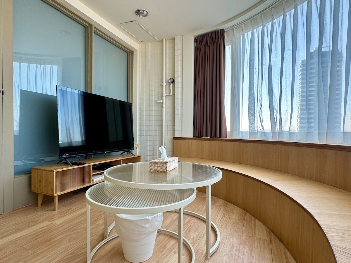 A bright living area features a large window with sheer curtains allowing natural light to fill the space. A modern television is positioned on a wooden stand, and a round coffee table is placed atop a warm wooden floor, complemented by circular seating arrangements.