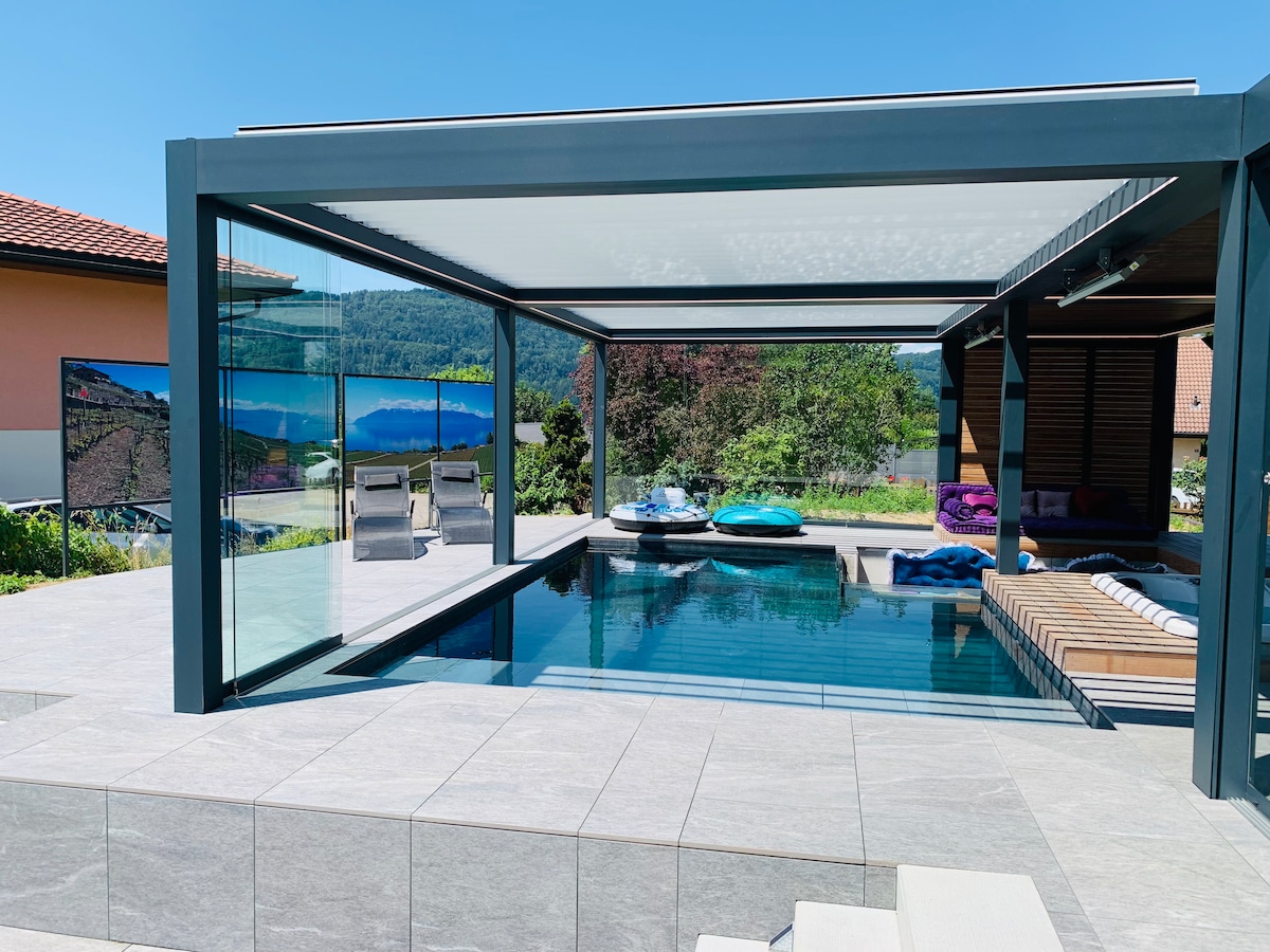 A contemporary pool area is enclosed by transparent walls, displaying a relaxing lounge with colorful cushions. The surface of the pool reflects the clear blue sky above, while a spacious deck is visible, furnished with minimalist seating. Lush greenery surrounds the space, enhancing the tranquil environment.
