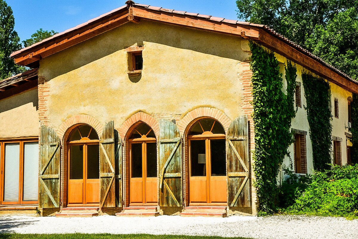 The exterior of the restored stables showcases a warm-toned facade adorned with climbing greenery. Wooden shutters frame the arched windows, allowing natural light to enter. A gravel pathway leads to the entrance, surrounded by lush foliage, contributing to the serene ambience.