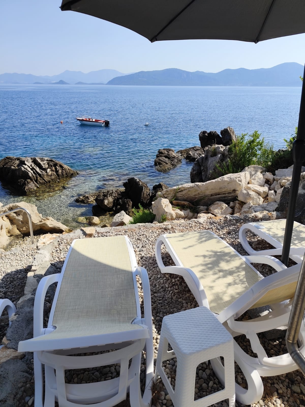 Sun loungers are arranged on a gravel area near the water's edge, offering a view of calm blue waters and distant islands. A small boat is visible on the water, while rocky formations are seen nearby, adding natural texture to the serene setting.