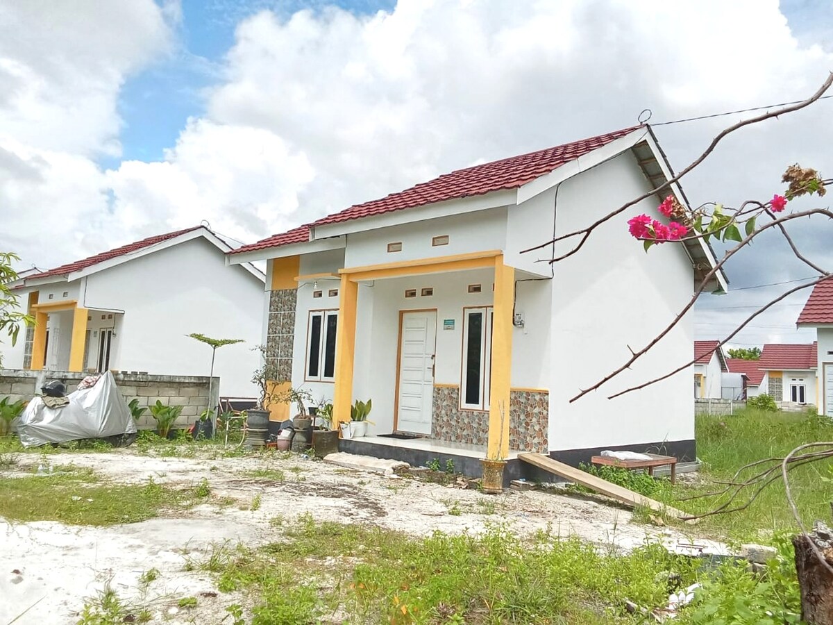 A single-story house with a simple design features a light-colored façade accented by colorful tilework around the entrance. The grassy area surrounding the house includes scattered plants and trees, with a distant view of neighboring homes under a partly cloudy sky.