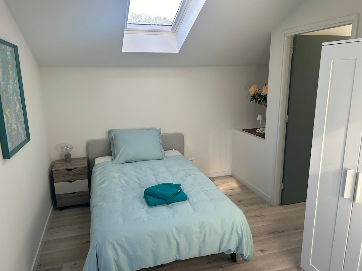A bright, cozy sleeping area features a single bed dressed in calming light blue linens. Natural light is allowed in through a skylight above. A small nightstand and a white wardrobe are positioned nearby, enhancing the organized space.