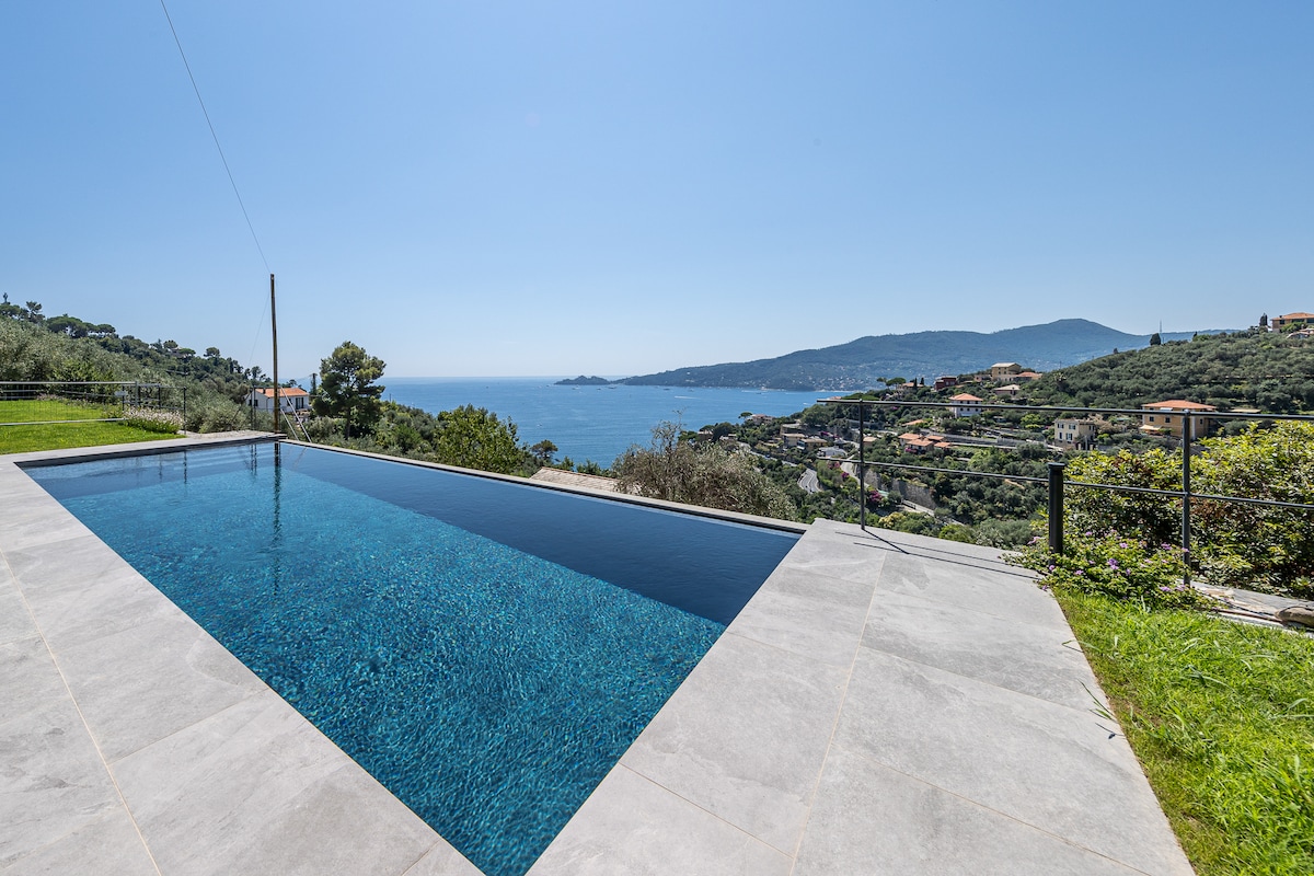 An infinity pool extends towards the horizon, offering clear blue water that reflects the surrounding landscape. Lush greenery borders the area, with views of the coastline and distant hills enhancing the serene atmosphere.