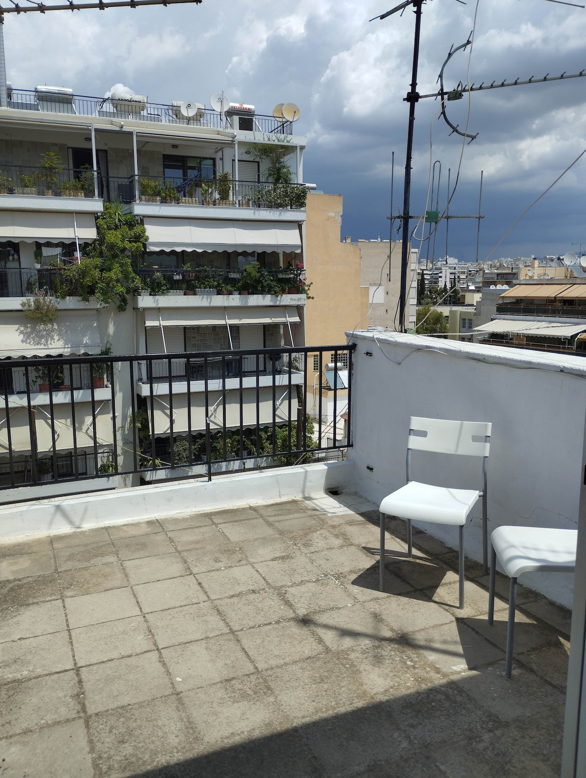 A private balcony features a simple setting, including a small table and two chairs. The area provides a view of nearby buildings adorned with greenery, while the open sky above creates a sense of spaciousness.