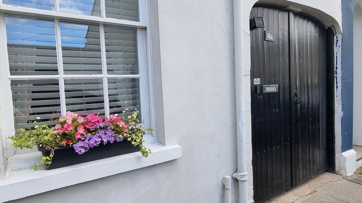 A charming exterior features a black door with a unique archway, framed by a white wall. A window with blinds offers a glimpse inside, while a vibrant flower box filled with pink and purple blooms adds a pop of color underneath.