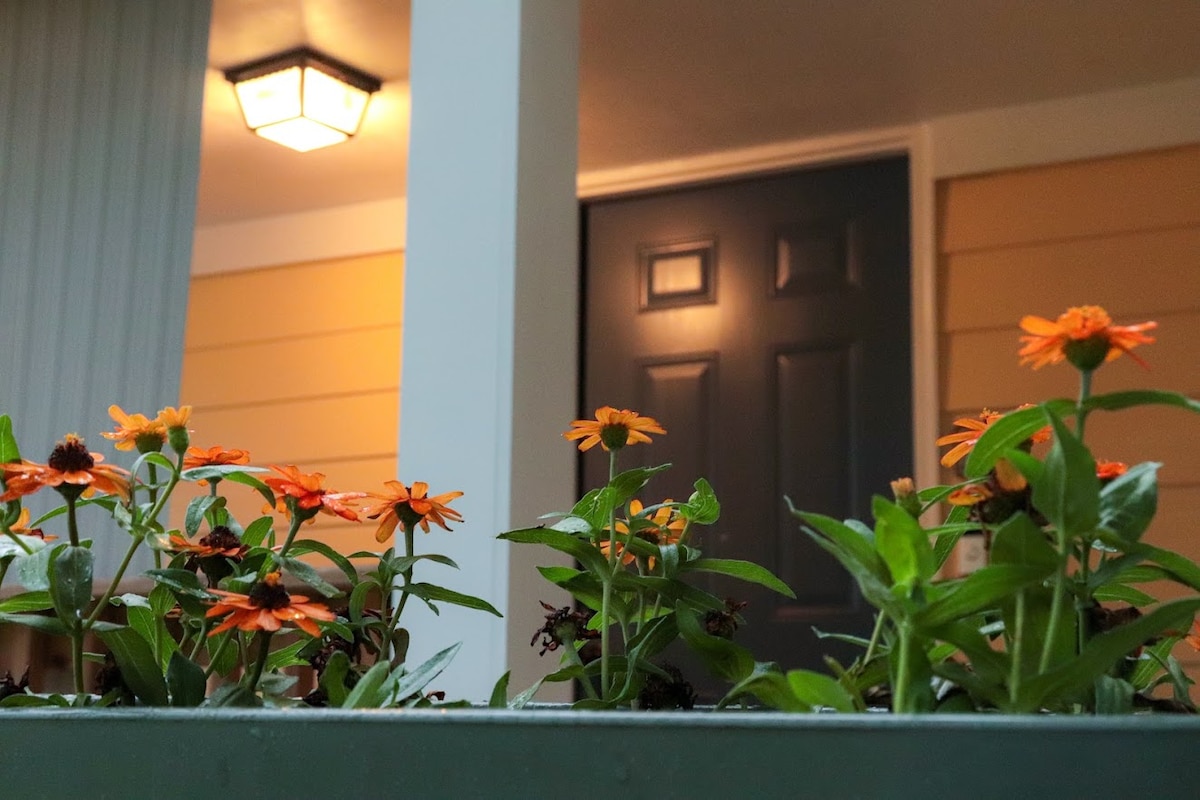 A welcoming entrance features a blue door framed by fresh flowers in vibrant orange and green. Soft lighting from a fixture above illuminates the area, creating a charming ambiance that invites guests to explore.
