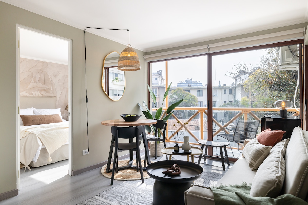 A bright living area features large windows allowing natural light to fill the space. A round dining table with black chairs is positioned near the window. A cozy sofa is complemented by decorative pillows. The bedroom is visible through the open door, showcasing a spacious bed.