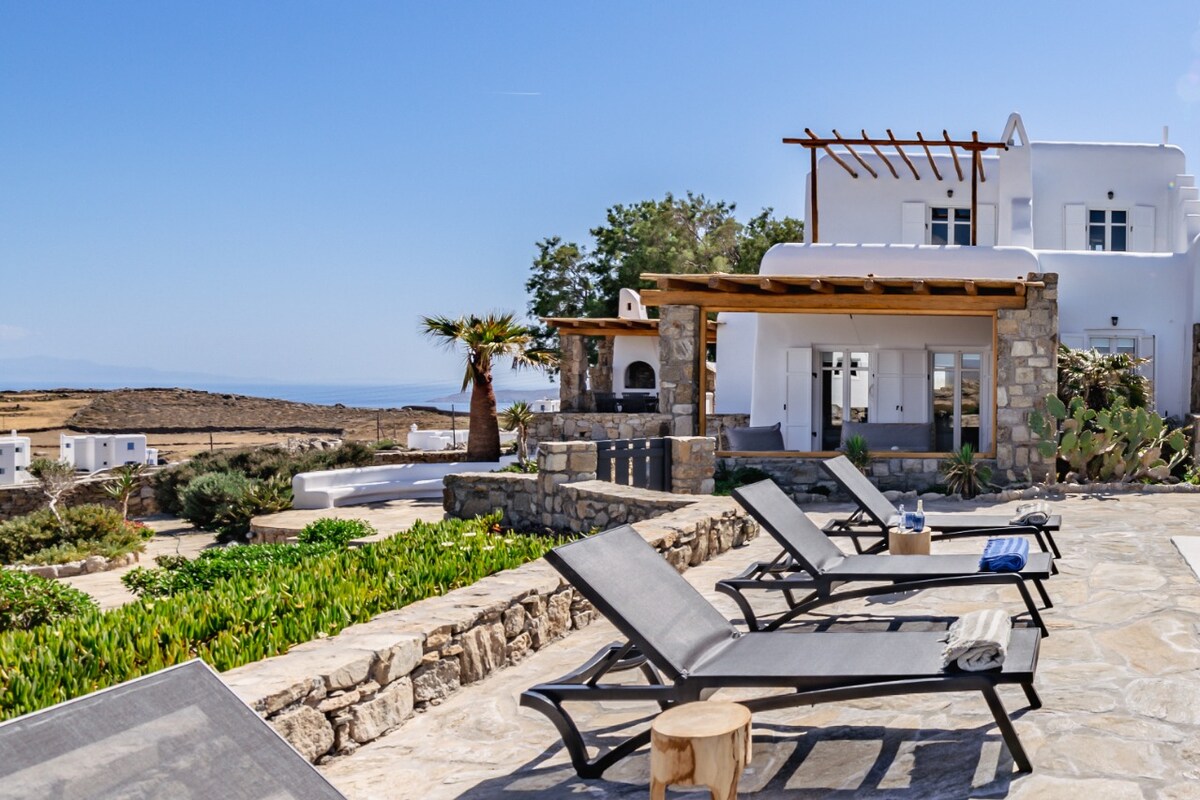 The outdoor terrace features several loungers arranged on a stone patio, surrounded by lush greenery and landscaping. A wooden pergola provides shade, and the villa's white exterior is visible in the background, complemented by a panoramic view of the ocean.