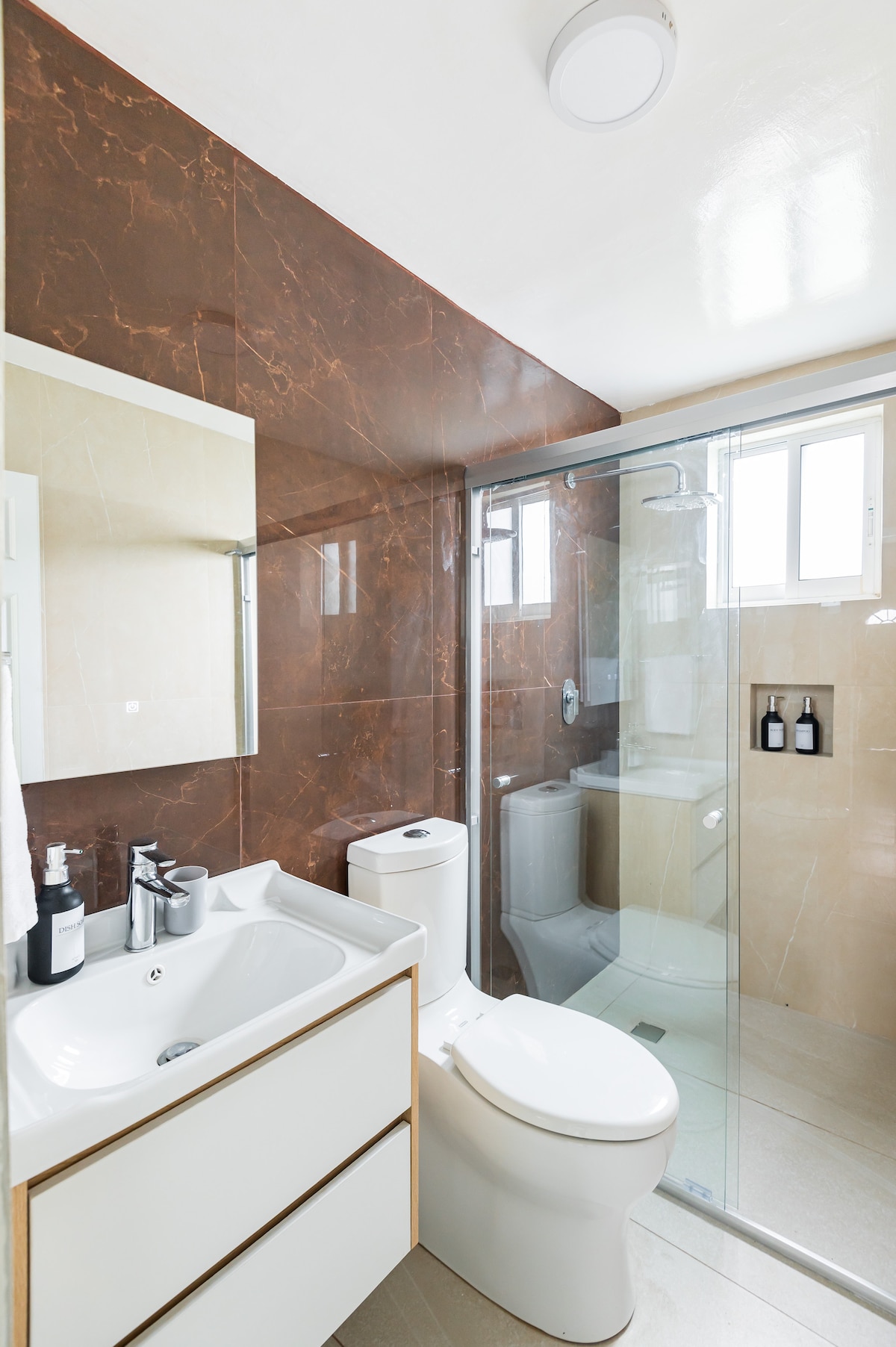 A well-appointed bathroom features modern fixtures, including a glass-enclosed shower and a contemporary vanity with storage. Earth-toned walls add warmth. Natural light filters through a nearby window, illuminating the space. Personal care products are arranged on the counter for convenience.