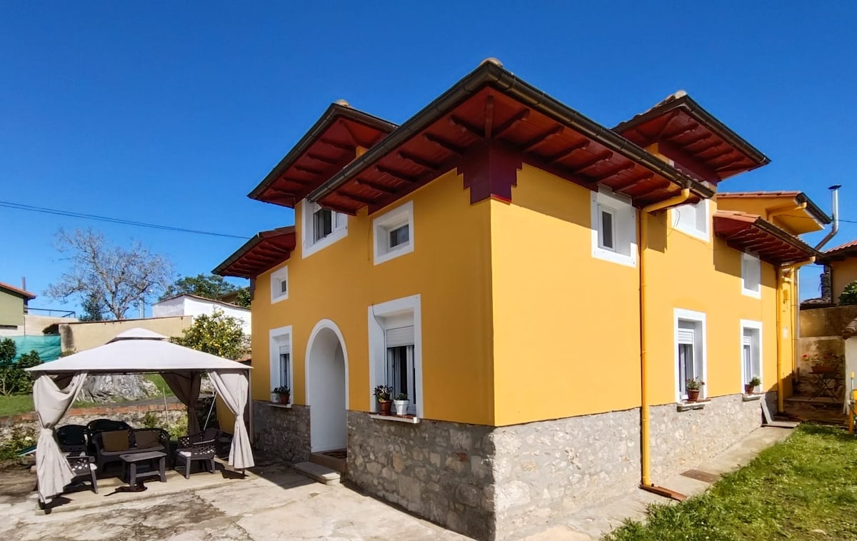 A vibrant yellow house stands under a clear blue sky, showcasing traditional architectural elements. A spacious outdoor area is adorned with a cozy gazebo, surrounded by well-maintained gardens that add to the inviting atmosphere of the property.