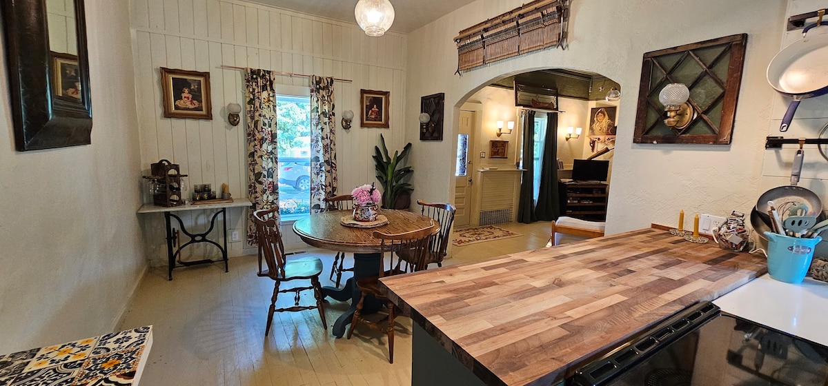 A warm and inviting kitchen area features a wooden dining table surrounded by several chairs. Decorative elements, including framed artwork and patterned curtains, add character. A spacious countertop is visible, complemented by a window view that allows natural light to enter the room.