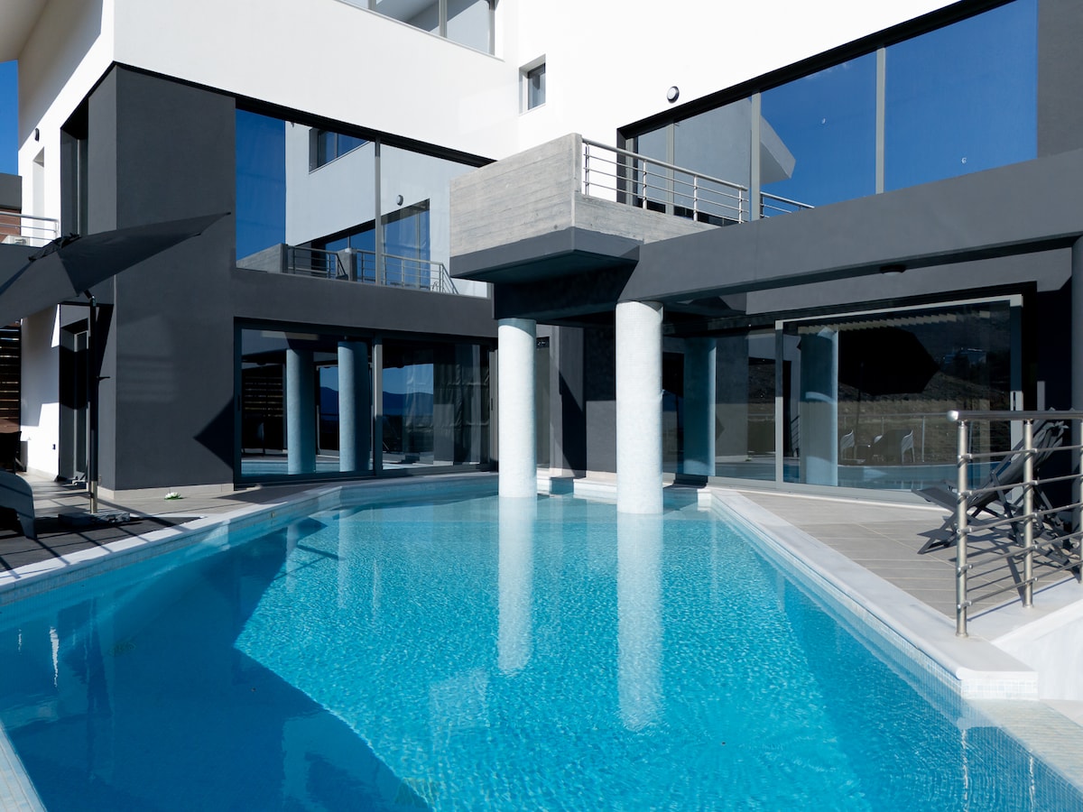 A modern villa exterior is showcased, featuring a private swimming pool surrounded by sleek concrete pillars and large glass windows. Reflections of the clear blue sky shimmer on the water's surface. Sun loungers are placed nearby under a large umbrella.