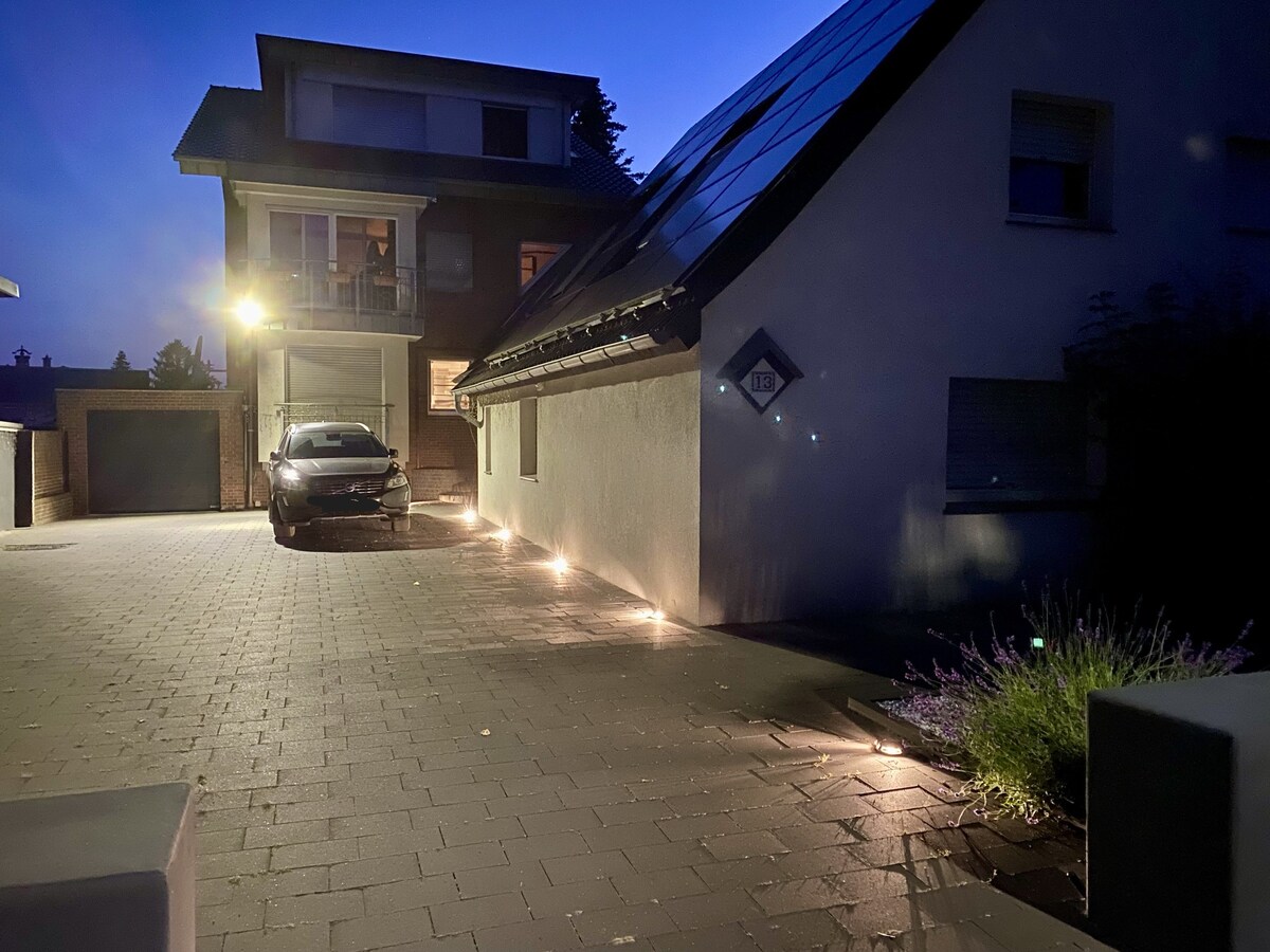 An exterior view of the double house is presented at dusk, revealing a driveway illuminated by soft lights. A car is parked to the side, while the structure features a modern design with solar panels on the roof and shrubs lining the edge.