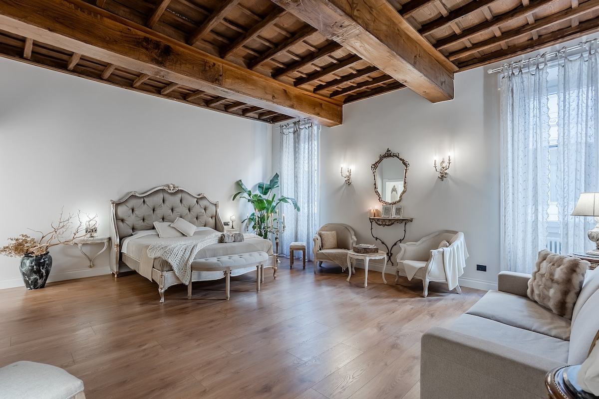 A spacious bedroom features a plush bed with a decorative headboard, complemented by an elegant seating area with armchairs. Large windows allow natural light to fill the room, enhancing the wooden ceiling beams and refined decor. A green plant adds a touch of nature to the ambiance.