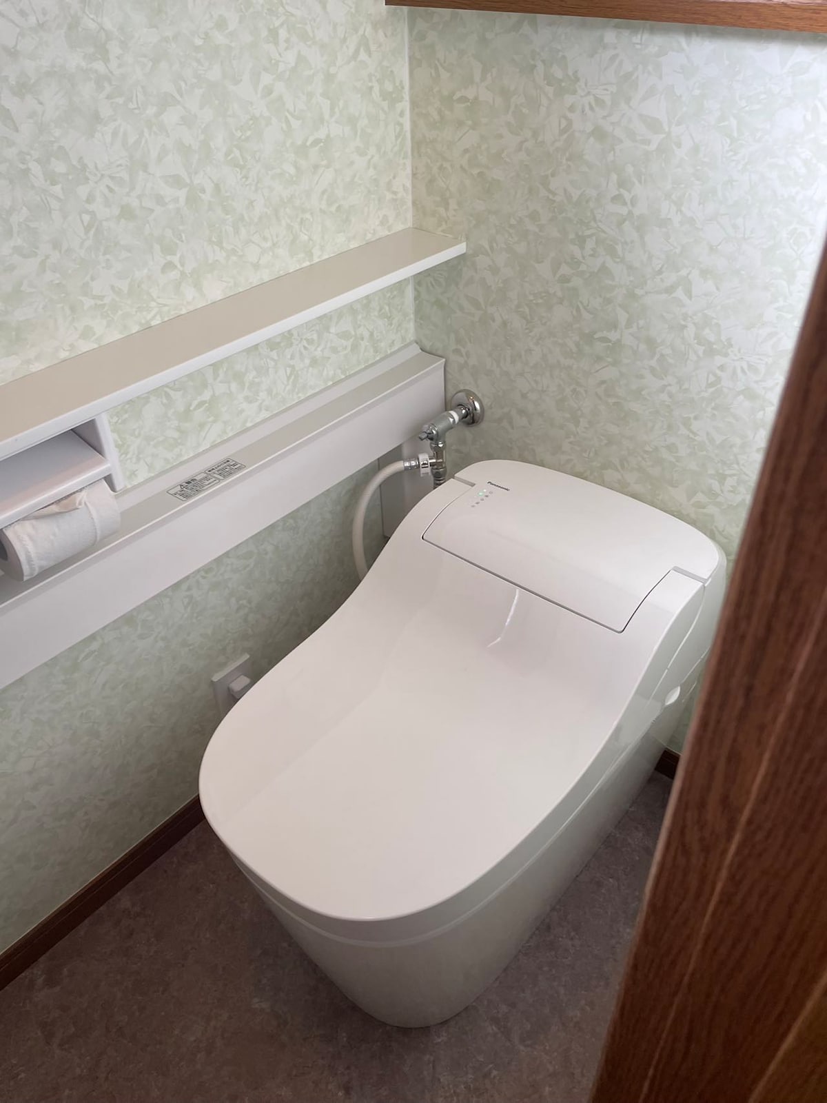 A modern toilet is situated in a spacious bathroom, featuring a sleek design and eco-friendly flush system. Light green patterned wallpaper complements the minimalistic aesthetic, while a shelf above provides space for essentials. The room is well-lit and maintained for comfort and cleanliness.