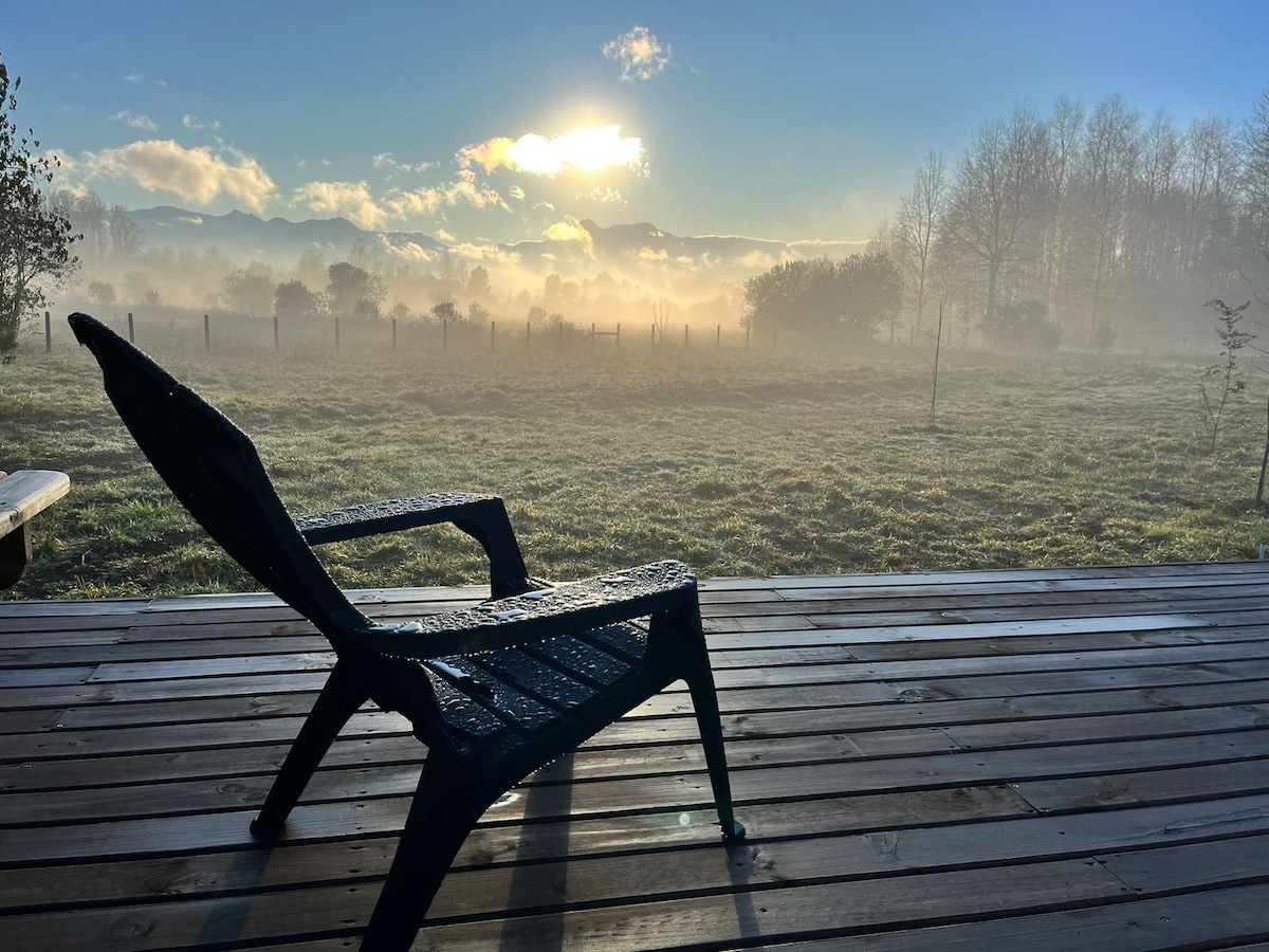 A black chair is positioned on a wooden deck, overlooking a misty landscape. Soft morning light filters through the fog, illuminating the fields and distant trees. The tranquil scene captures the atmosphere of the surrounding natural beauty.
