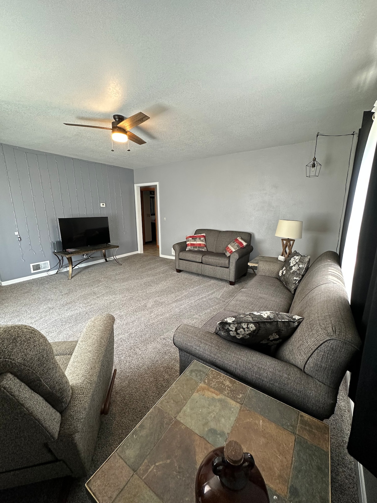 A spacious living area is shown with neutral-toned walls and carpet. A ceiling fan provides comfort overhead. A flat-screen television is positioned on one wall, while two sofas with patterned cushions and a side table create a welcoming seating arrangement.