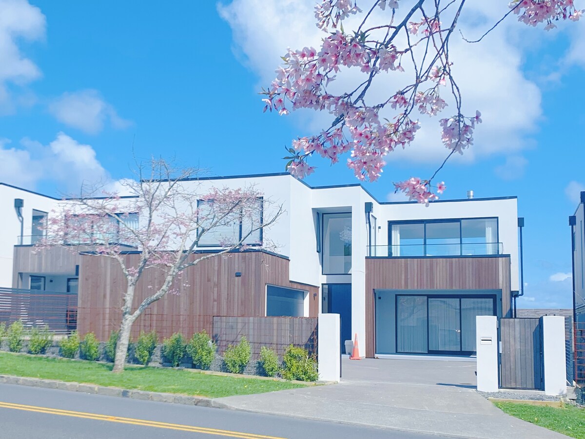 A modern minimalist home is presented with clean lines and a fresh facade, complemented by natural wood accents. Lush greenery and flowering branches frame the entrance, while large windows reflect the blue sky, enhancing the open and airy feel of the property.