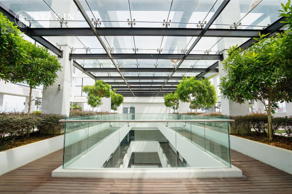An open communal space is viewed, featuring a modern design with a glass ceiling that allows natural light to flood in. Neatly arranged trees provide greenery, enhancing the tranquil atmosphere. The wooden flooring adds warmth, creating a harmonious environment for relaxation.