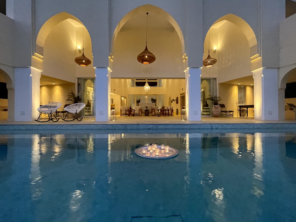 A serene pool area is illuminated by soft lighting, reflecting the gentle glow of candles floating on the water. Arched openings frame a spacious communal area, where elegant furnishings and decorative lamps create a calming ambiance.