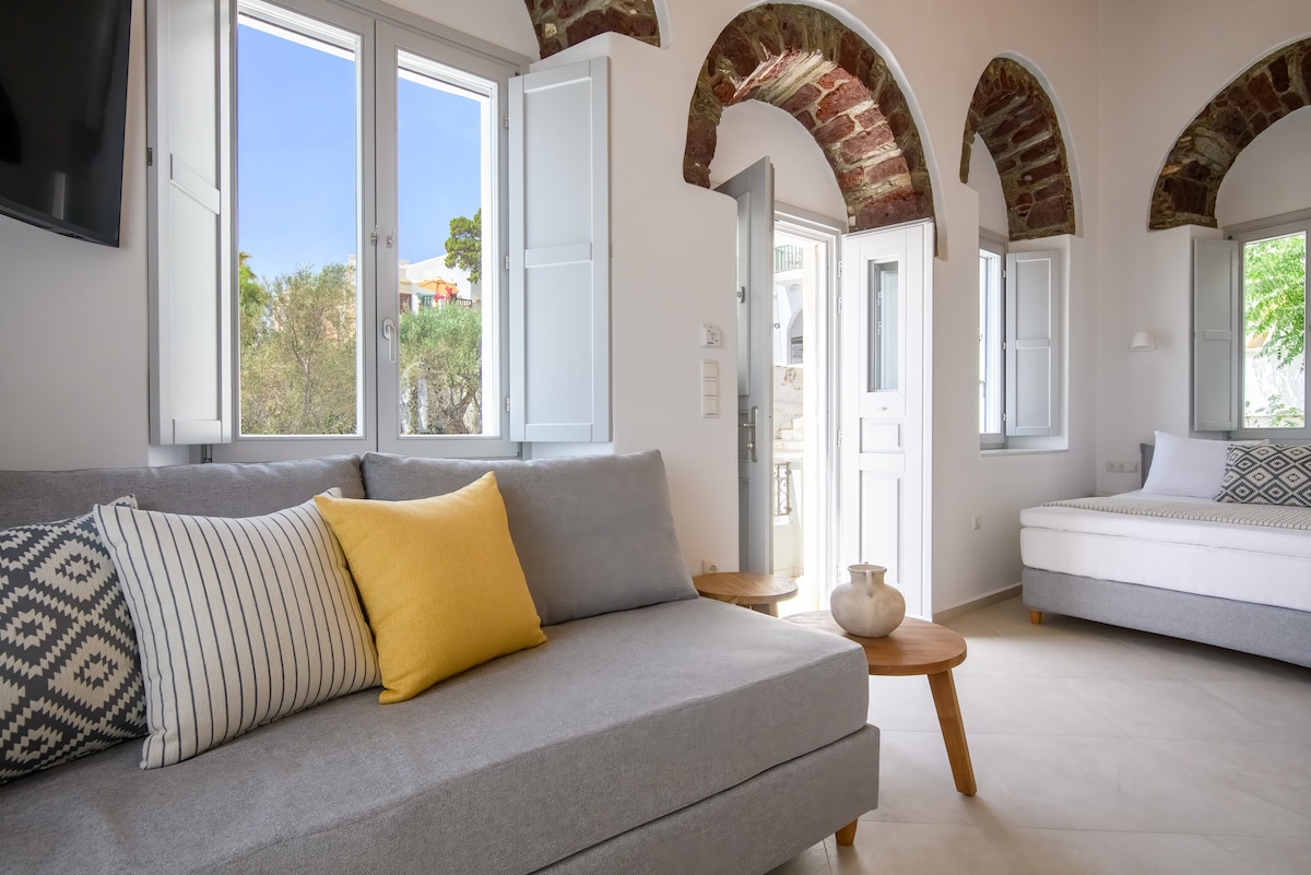 A contemporary sitting area features a grey sofa with decorative cushions. Natural light fills the space through arched windows, framed by traditional shutters. A light wooden table is positioned nearby, complementing the soft neutral tones of the interior.