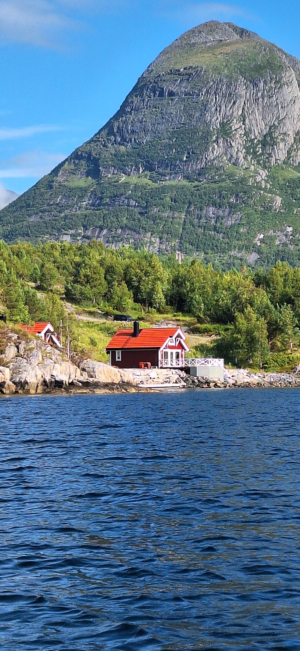 A charming red cabin is positioned along the water's edge, surrounded by lush greenery and mountainous terrain. The cabin features a white-roofed balcony that overlooks the serene blue waters, reflecting the natural beauty of the area.