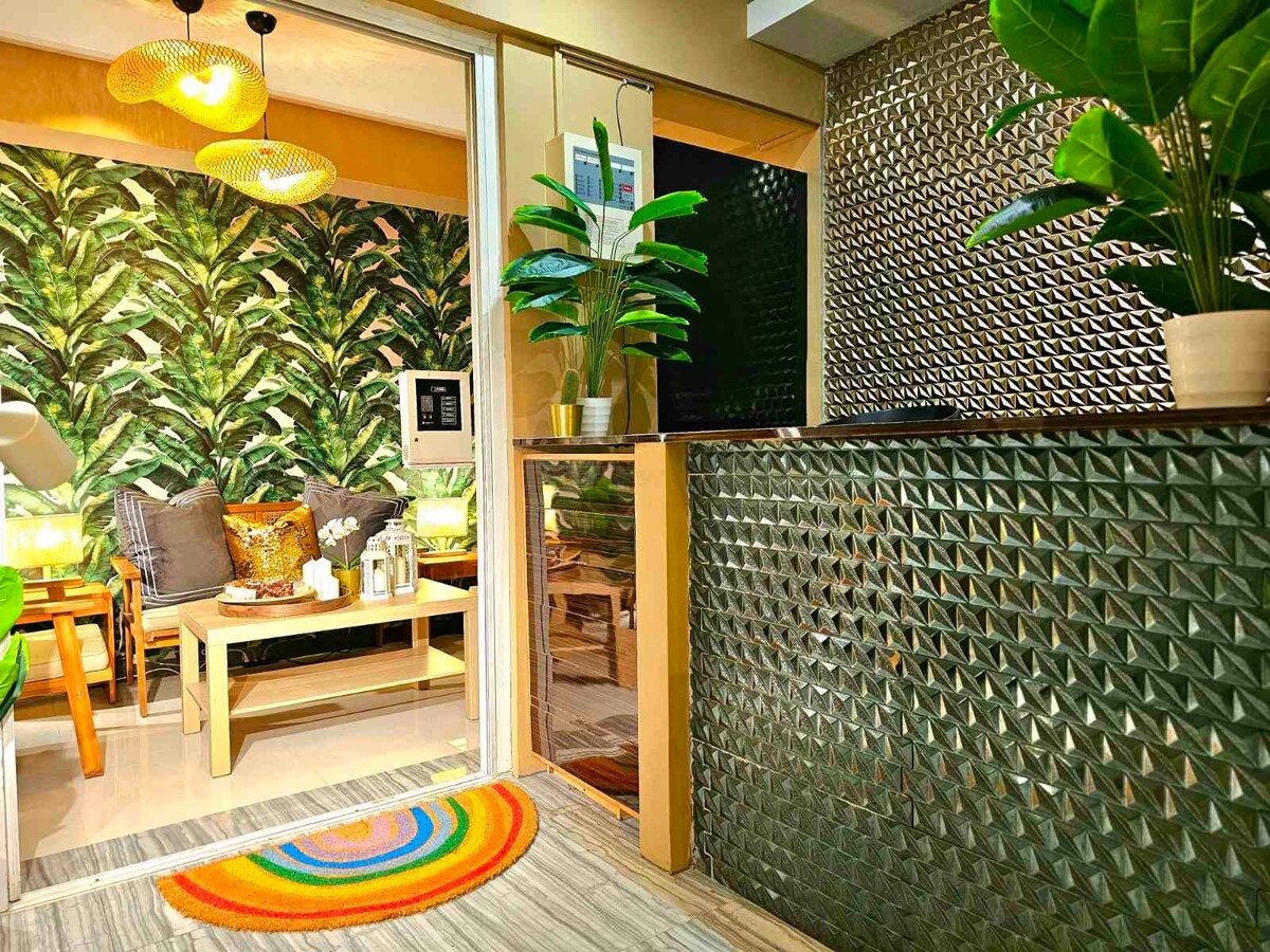 A welcoming entryway features a textured wall with reflective tiles. A circular, multicolored rug is placed at the entrance. In the background, a cozy seating area showcases vibrant green foliage wallpaper, complemented by a light wooden table and decorative cushions.