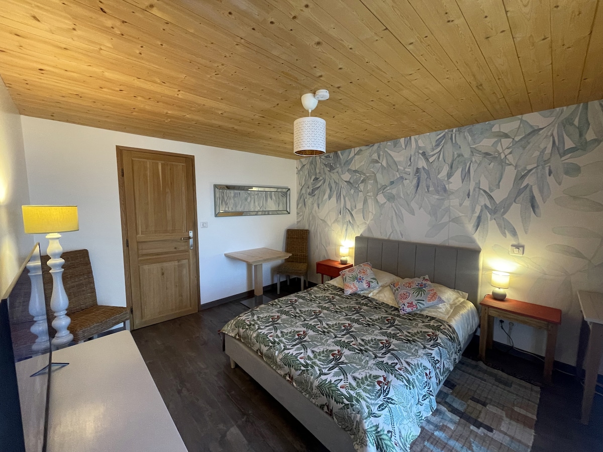 A spacious room features a large bed with a floral-patterned duvet and decorative pillows. Flanking the bed are two red side tables, and a wooden desk and chairs are positioned nearby. The ceiling showcases natural wood paneling, and a mirror reflects the light from the lamp.