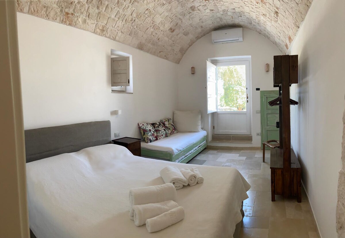 The sleeping area features a spacious bed with crisp white linens and two stacked single beds that serve as a couch. Natural light filters through a window, illuminating the room's arched stone ceiling and neutral-toned walls, with a wooden wardrobe and folding rack adding functionality.
