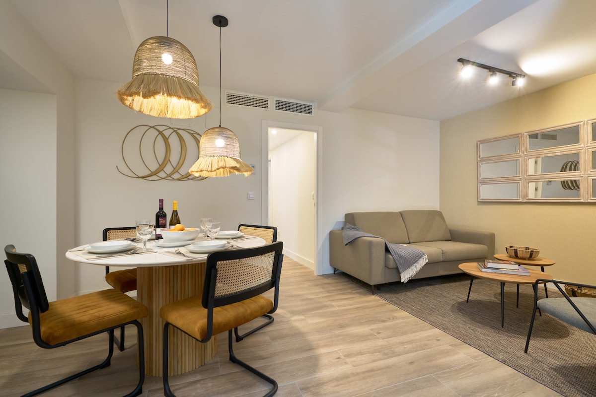 A modern dining area is featured, showcasing a round table with a textured surface and four black chairs. Two pendant lights hang above, providing warm illumination. A sectional sofa is positioned nearby, and a stylish coffee table complements the space. Decorative elements include wall art and a neutral color palette.