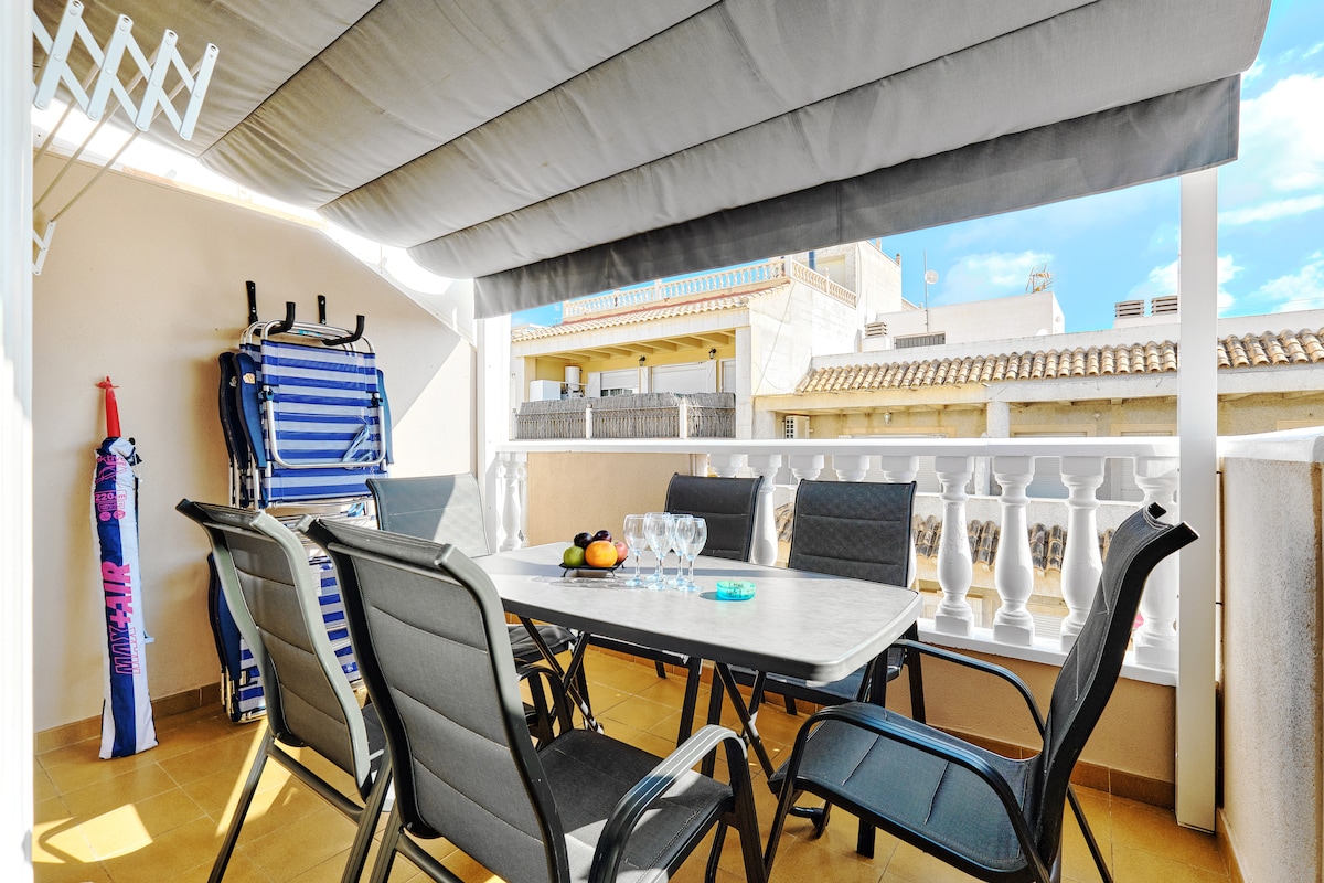 A spacious outdoor terrace is featured, showcasing a large table surrounded by six black chairs. Sun loungers are positioned nearby, and a sunshade provides protection from the sun. The background reveals additional buildings and rooftops under a bright blue sky.