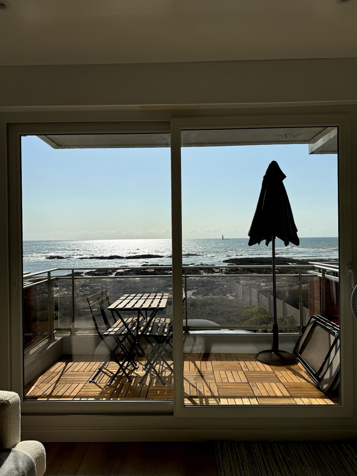 A sliding glass door frames a view of the ocean, reflecting sunlight across the water. A balcony is visible, featuring a small dining table and two chairs, alongside a sunshade. The serene atmosphere invites relaxation and outdoor enjoyment.