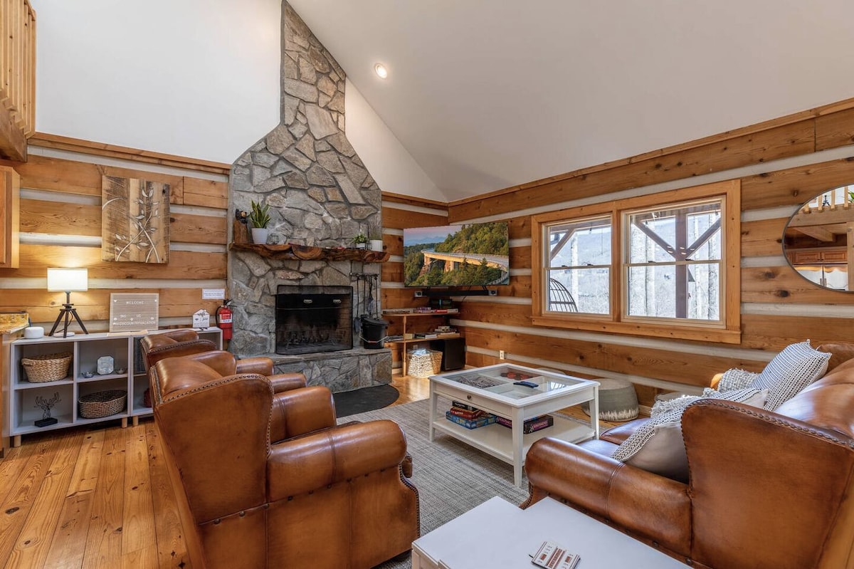 A cozy living room features a stone fireplace with a wooden mantle, surrounded by rustic wooden walls. Comfortable leather armchairs are positioned near a central coffee table, while large windows allow natural light to fill the space, showcasing the view outside.