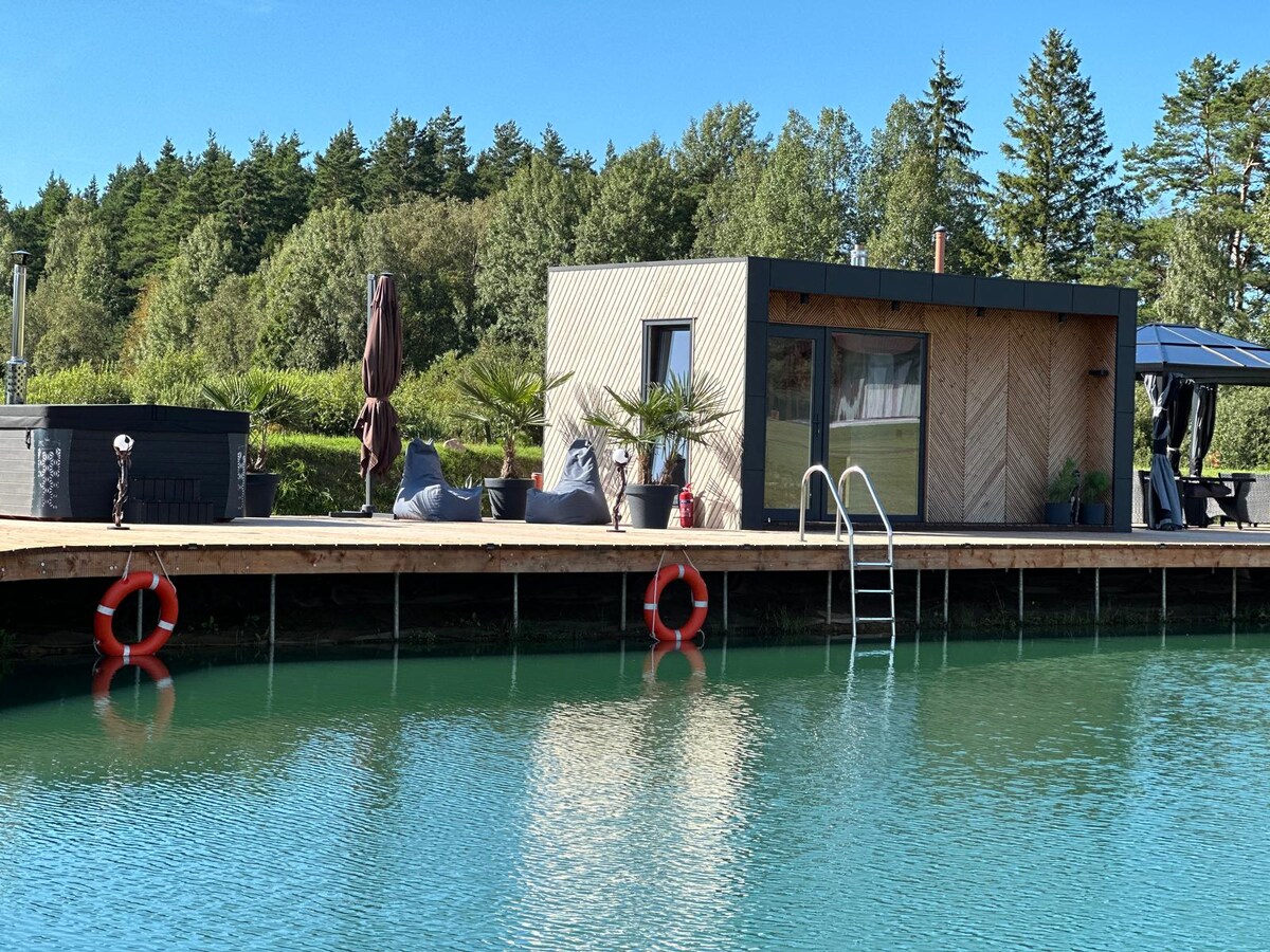 A modern cabin is situated on a dock by a serene pond, surrounded by lush trees. The cabin features large windows and a patio area, with lounge chairs and a hot tub visible. Colorful life rings float nearby, enhancing the tranquil waterfront setting.
