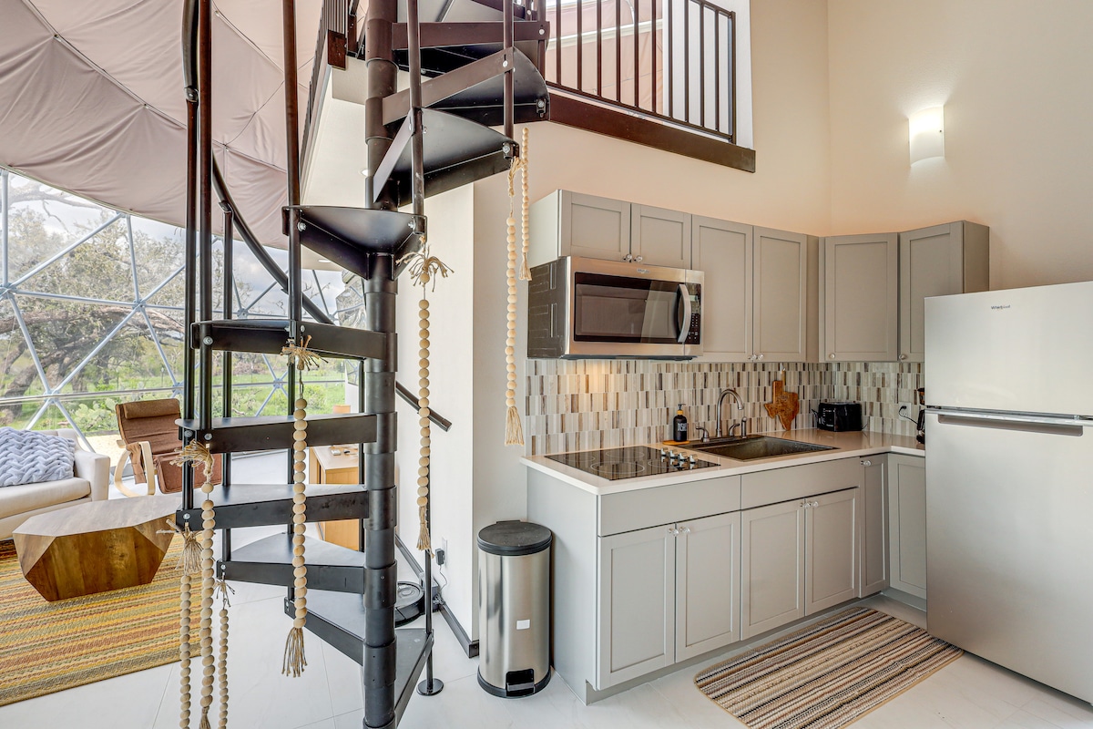 An open kitchen area features modern appliances, including a microwave and refrigerator, along with gray cabinetry and a stylish backsplash. A spiral staircase leads to an upper loft space, while natural light floods the room through a large window, enhancing the airy feel.