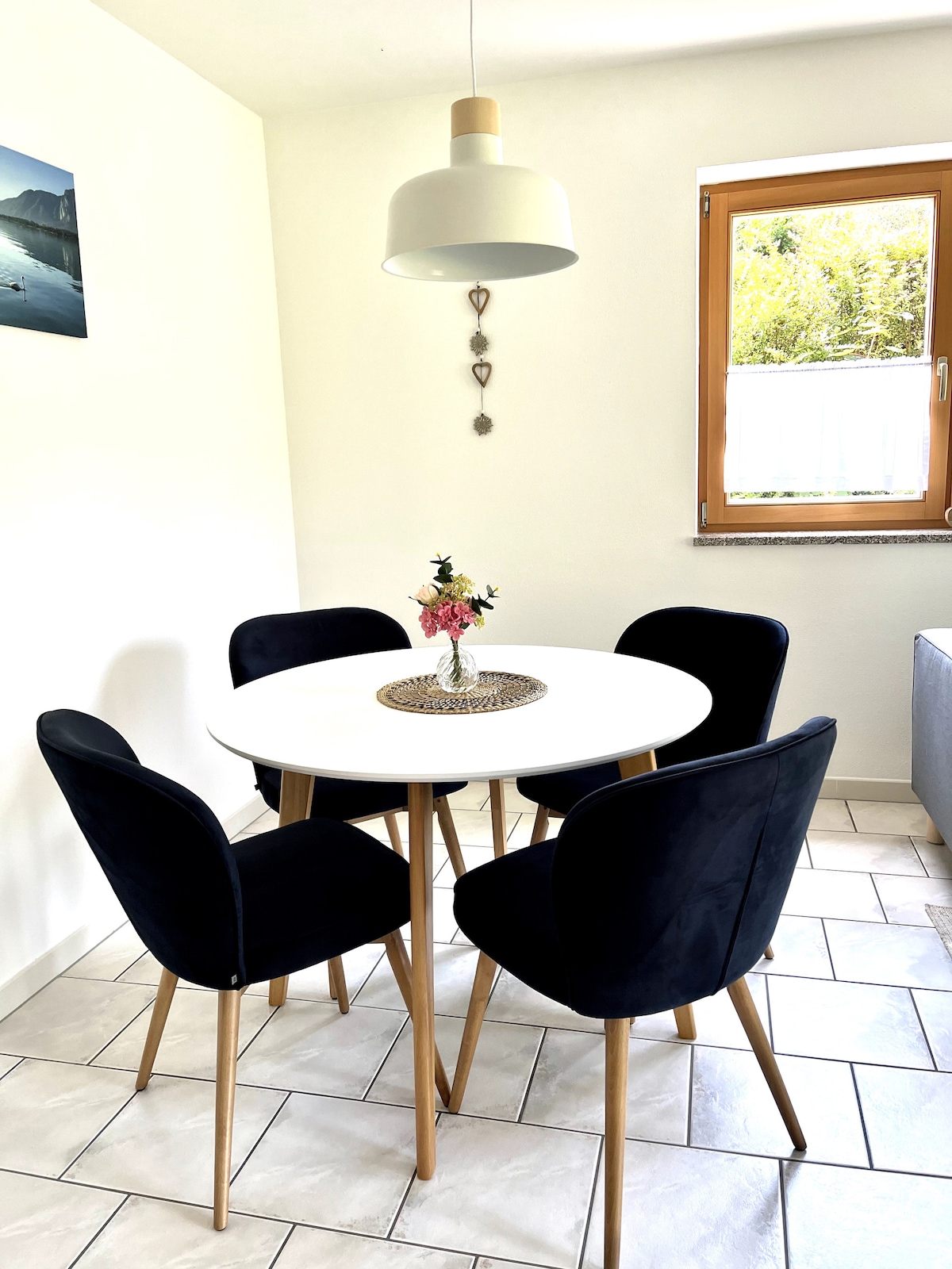 A round dining table is surrounded by four upholstered chairs, creating a cozy dining area. A small vase with flowers sits at the center of the table. Natural light enters through a window adorned with light-colored curtains, adding warmth to the space.