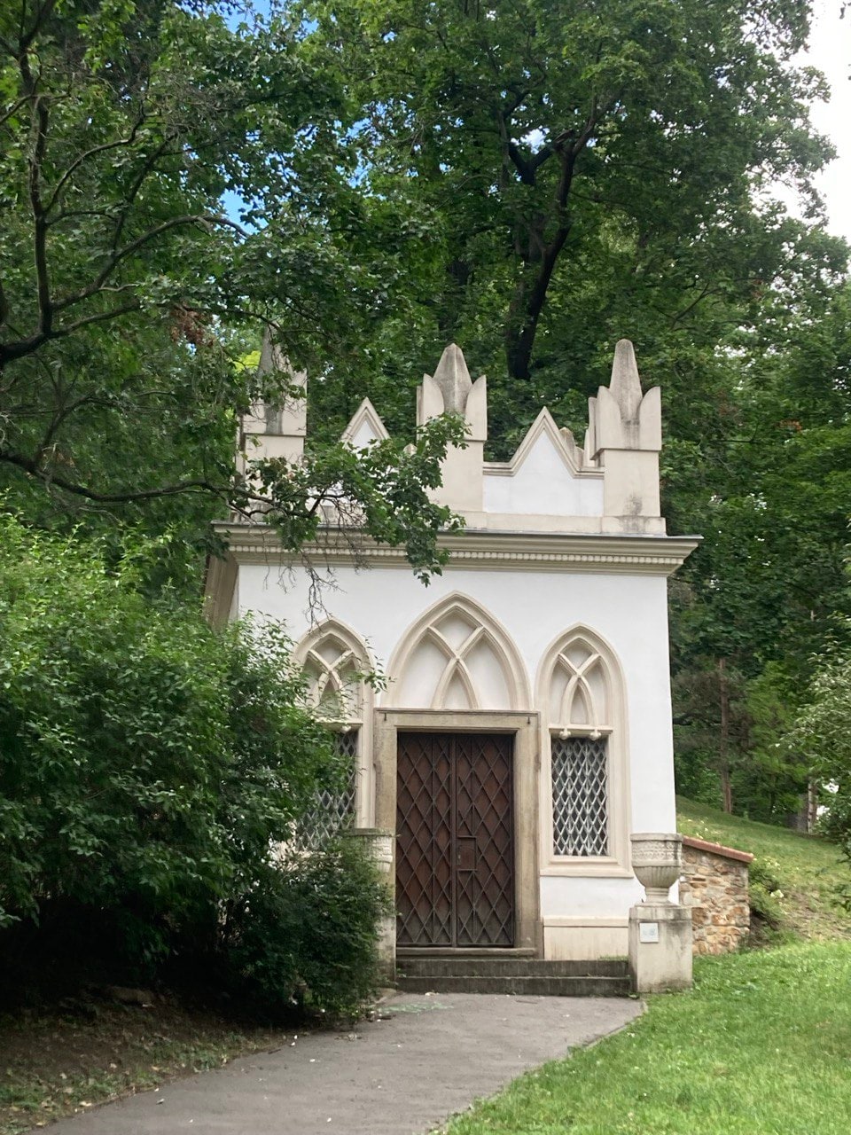 A small white structure with pointed arches and decorative elements stands surrounded by lush greenery. The building features a wooden door with intricate details and lattice windows, set against a backdrop of tall trees and a well-maintained path leading up to it.