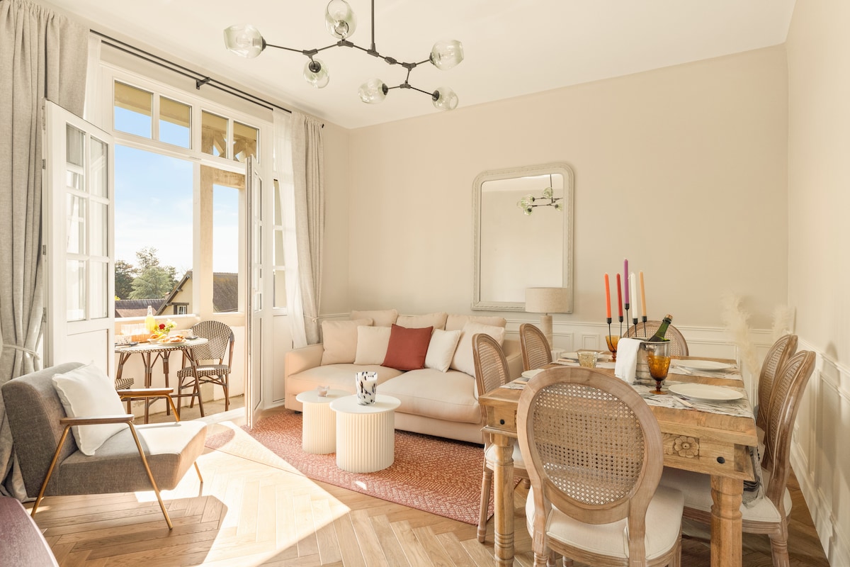 A bright dining and living area features a light-colored sofa accompanied by a round coffee table. A wooden dining table is surrounded by stylish chairs. Large windows allow natural light to fill the space, creating an open and serene atmosphere. A neutral color palette enhances the room's charm.