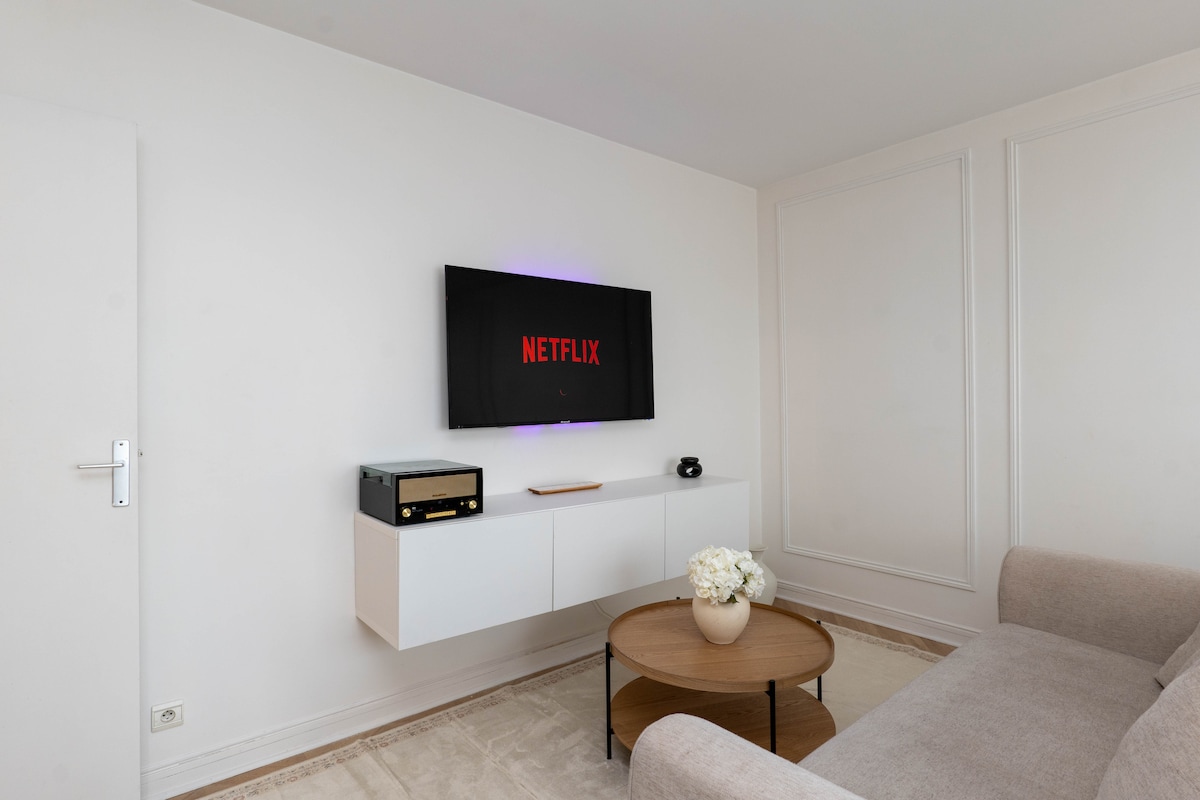 The living area features a wall-mounted television displaying the Netflix logo, accompanied by a modern white storage unit below. A round coffee table sits in front of a comfortable sofa, and a decorative vase with flowers is positioned on the table.