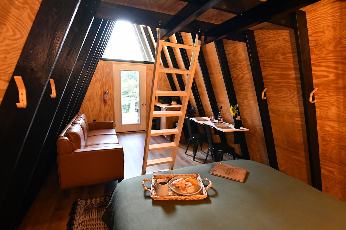 An inviting A-frame interior features wooden walls and a cozy queen bed. A wooden ladder leads to a loft area. A comfortable sofa is positioned next to a dining table for two, while a door provides access to the outdoors.