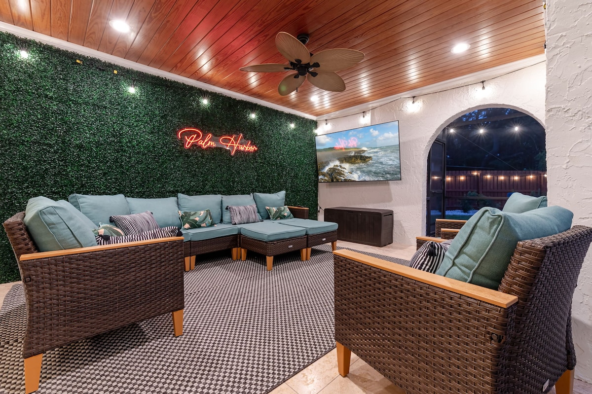 A comfortable outdoor seating area is presented with plush, turquoise cushions on dark wicker furniture. The space is enhanced by artificial greenery on the walls and features a large flat-screen TV, providing a relaxing environment for social gatherings.