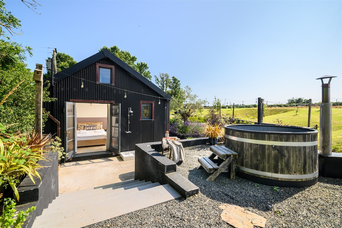 An outdoor view of The Hiding Place highlights a black wooden structure, blending naturally with its surroundings. A wood-fired hot tub is positioned nearby, accompanied by a stone seating area. Large bifold doors open to reveal a glimpse of the interior space, surrounded by lush greenery.