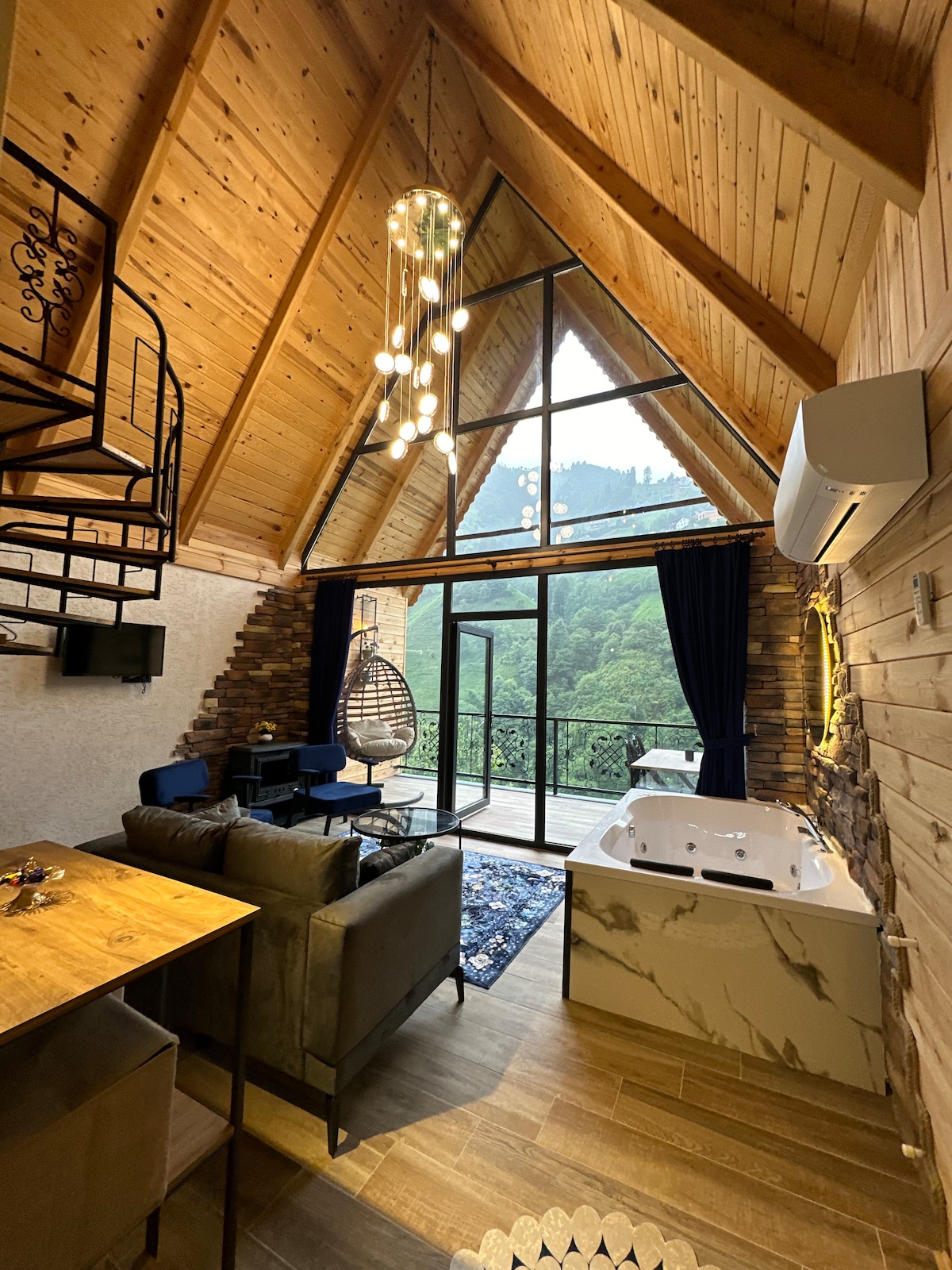 The interior features a high-ceilinged space with wooden accents and large windows overlooking a lush mountainous landscape. A cozy seating area includes a dark sofa and armchairs, while a stylish dining table and a jacuzzi are also present. A spiral staircase leads to an upper level.
