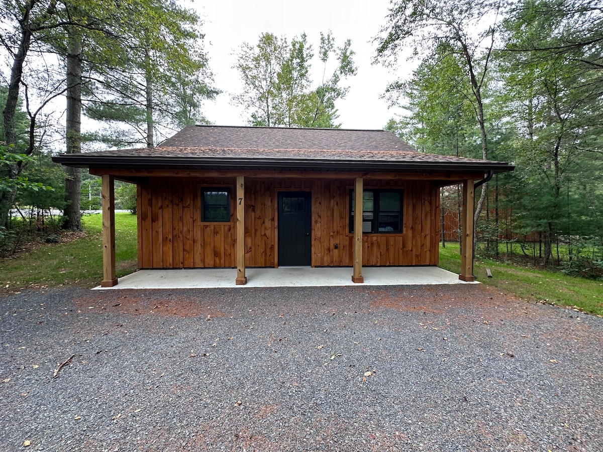A wooden cabin with a covered porch sits nestled among trees. The exterior features natural wood siding and a dark front door. Gravel lines the path in front of the cabin, and a spacious outdoor area is visible, surrounded by greenery.