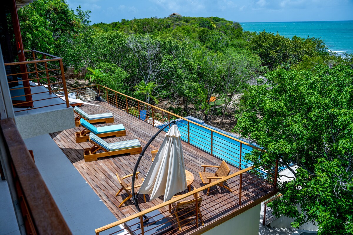 A spacious outdoor deck is visible, featuring comfortable lounge chairs and a stylish hanging chair. A pool is situated nearby, with lush greenery surrounding the area and offering views of the ocean in the distance.