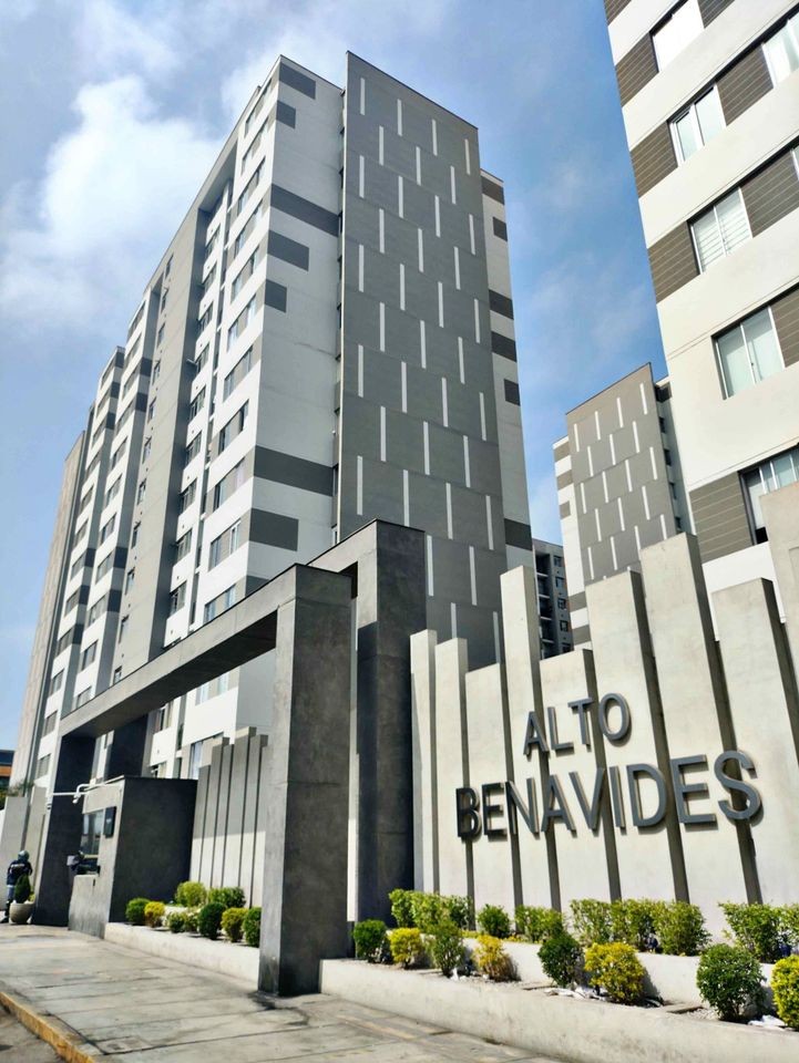 A modern apartment building is showcased, featuring a sleek façade with geometric patterns. The entrance is marked by a concrete wall displaying the name 'ALTO BENAVIDES,' framed by neatly trimmed shrubs along the pathway and a clear blue sky above.