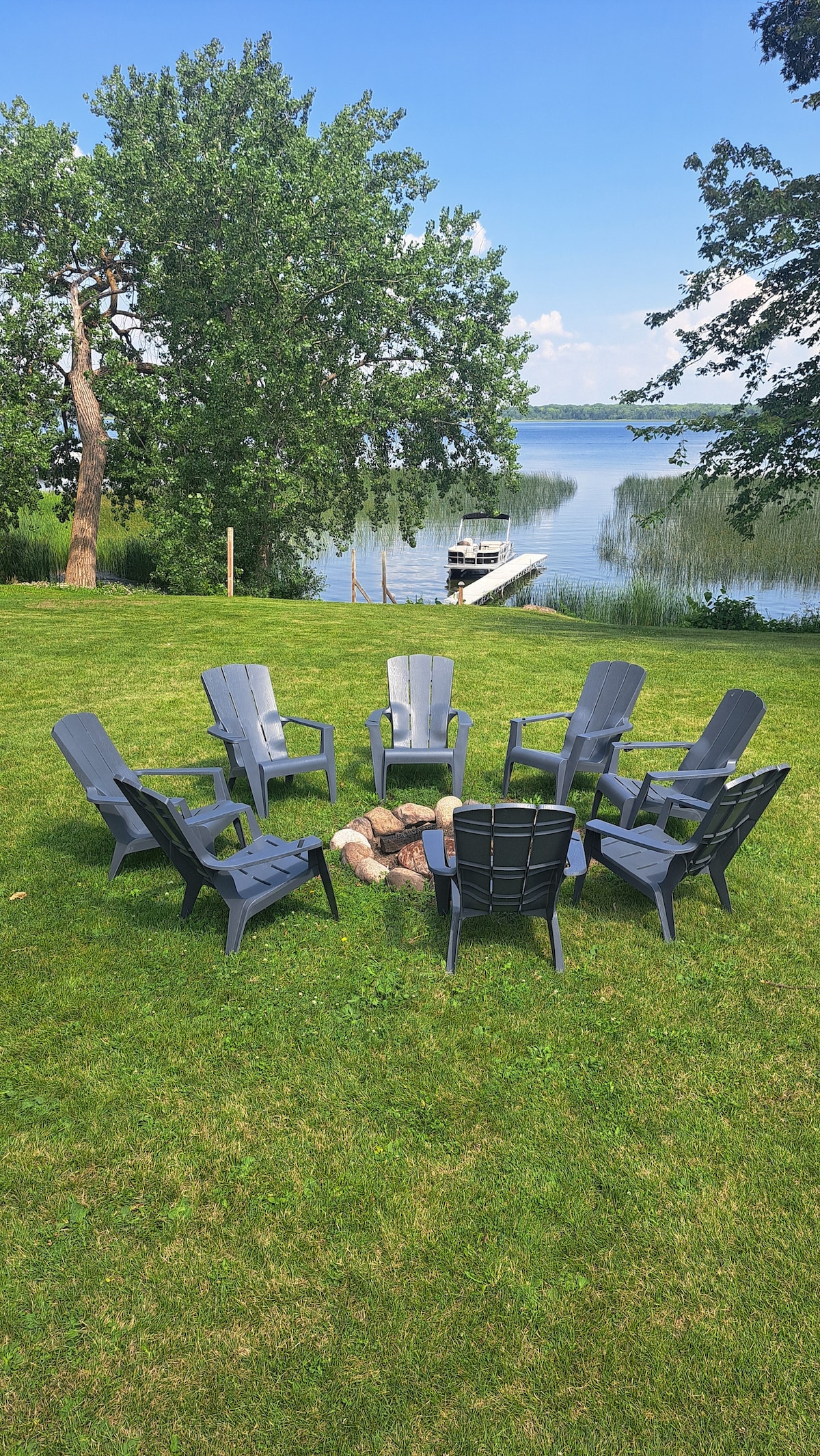 A circular arrangement of dark chairs surrounds a fire pit on a grassy lawn. The serene lake is visible in the background, framed by trees under a clear blue sky. A boat is docked in the distance, suggesting easy access to water activities.