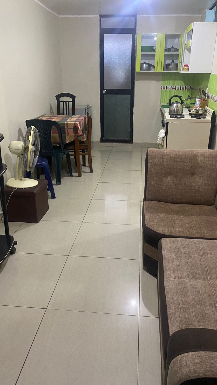 A clean and functional kitchen area is visible, featuring green cabinets and essential appliances. A dining table with four chairs is arranged nearby. A cozy seating area with a two-tone couch and a standing fan is positioned to the side, enhancing the overall spaciousness.