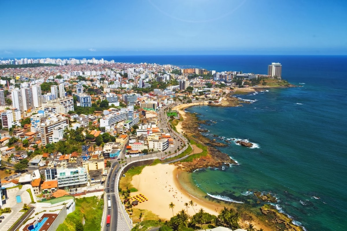 An aerial view showcases the coastline and urban landscape of Rio Vermelho, featuring sandy beaches, rocky outcrops, and a vibrant array of buildings. The blue ocean contrasts beautifully with the cityscape, while palm trees line the shore, adding to the coastal charm of the area.