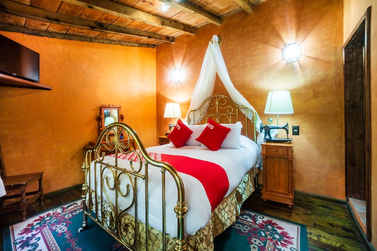 A cozy room is highlighted by warm orange walls and wooden beams. A decorative gold bed frame supports a soft white comforter, accented with red pillows. A rug lies on the hardwood floor, and bedside lamps illuminate the space for a welcoming feel.
